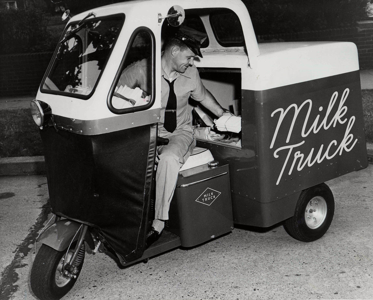 A vintage black and white photo of a milk truck driver sitting in a small three-wheeled milk delivery vehicle with 'Milk Truck' written on the side.